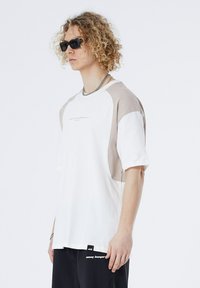 Short-sleeved t-shirt in white with beige raglan sleeves, featuring a logo in the center and a textured fabric detail on the sides.