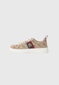 Beige canvas-sneakers med texturerat logomönster, vit gummisula och broderad patch med en krona och färgglada accent.
