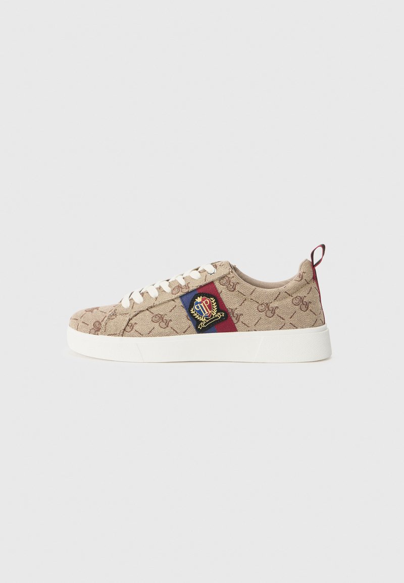 Beige canvas-sneakers med texturerat logomönster, vit gummisula och broderad patch med en krona och färgglada accent.