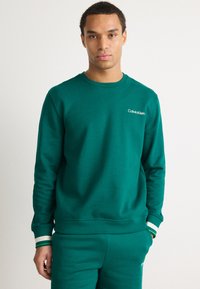 Zelená bavlněná mikina se zaobleným výstřihem, dlouhými rukávy a bílým logem Calvin Klein. Obsahuje žebrované manžety se zelenými a bílými pruhy.