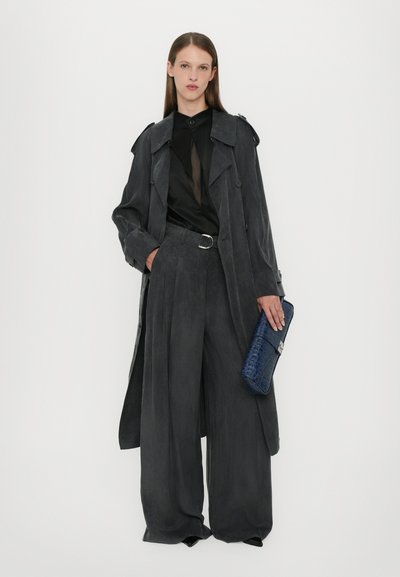 Grijze trenchcoat met een losse pasvorm, gecombineerd met grijze broek met wijde pijpen. Zwart top eronder, met een gestructureerde blauwe clutch vast.