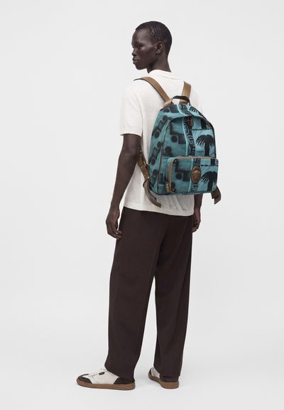 Homme portant une chemise blanche à manches courtes et un pantalon foncé, portant un sac à dos à motif bleu sarcelle et noir avec des sangles beige, debout devant un fond uni.