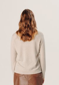 Beiger Pullover mit gerippten Bündchen und Saum, glatter Textur und klassischer Passform. Getragen mit einem braunen Lederrock, von hinten zu sehen.