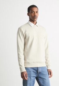 Sudadera beige con el logo "BOSS" en relieve, combinada con una camisa blanca de cuello. Textura suave, puños acanalados y corte casual. Jeans de denim finos.