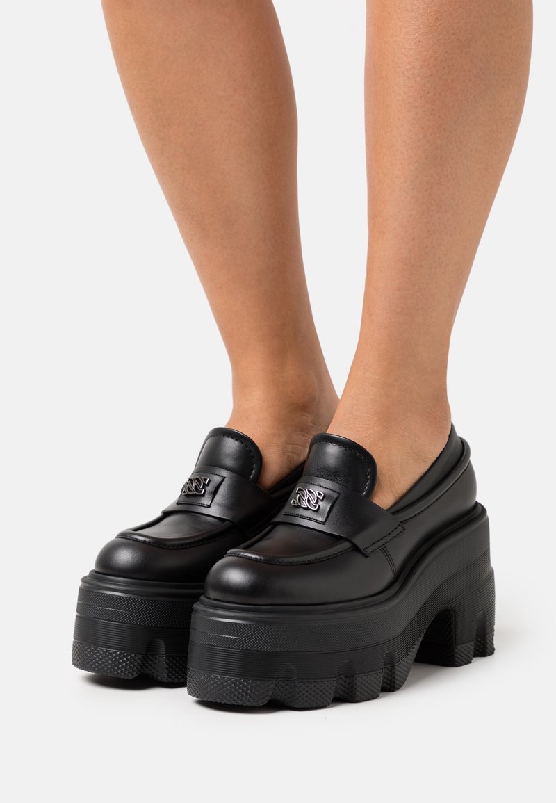 Mocassins à plateforme noires avec une tige en cuir lisse, une semelle épaisse et structurée, et un accent métallique sur le devant. Motif de bande de roulement en caoutchouc sur la semelle.