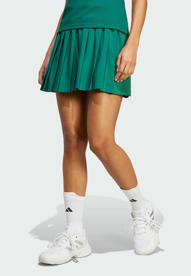 Grüner plissierter Tennisrock, kombiniert mit weißen Sportschuhen und Socken, der eine glatte Stofftextur und eine ausgestellte Silhouette zeigt.