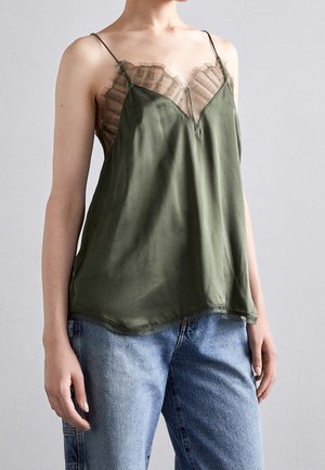 Kvinde iført en ærmeløs olivengrøn camisole med gennemsigtige stribede blonder, kombineret med lyseblå denimjeans.