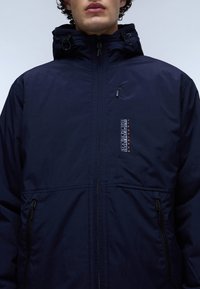 Navykleurige jas met een ritssluiting, een trekkoordcapuchon, zijzakken en een logo op de borst. Gladde, matte stoftextuur.