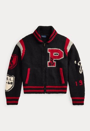 Kvinde med flettet hår iført sort og rød varsity sweater, grå bukser, hvid rullekrave, der holder en sort håndtaske mod en neutral baggrund.