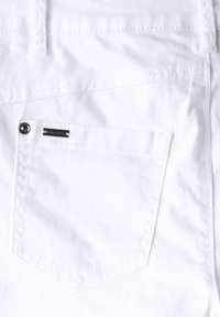 Primer plano de tejido de mezclilla blanca que presenta una textura suave, un bolsillo trasero con un botón y una etiqueta de marca negra adjunta.
