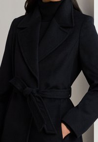 Donna che indossa un cappotto di lana nero con cintura sopra un dolcevita nero, con una mano nella tasca del cappotto.