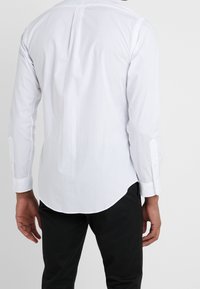 Mężczyzna w białej koszuli z długim rękawem i czarnych spodniach, pokazany od tyłu na jednolitym tle.