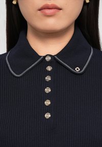 Tume ribaga kaelusega polo, millel on valged ääred, kuus dekoratiivset hõbedast nuppu ja väike lillemuster kaeluses.