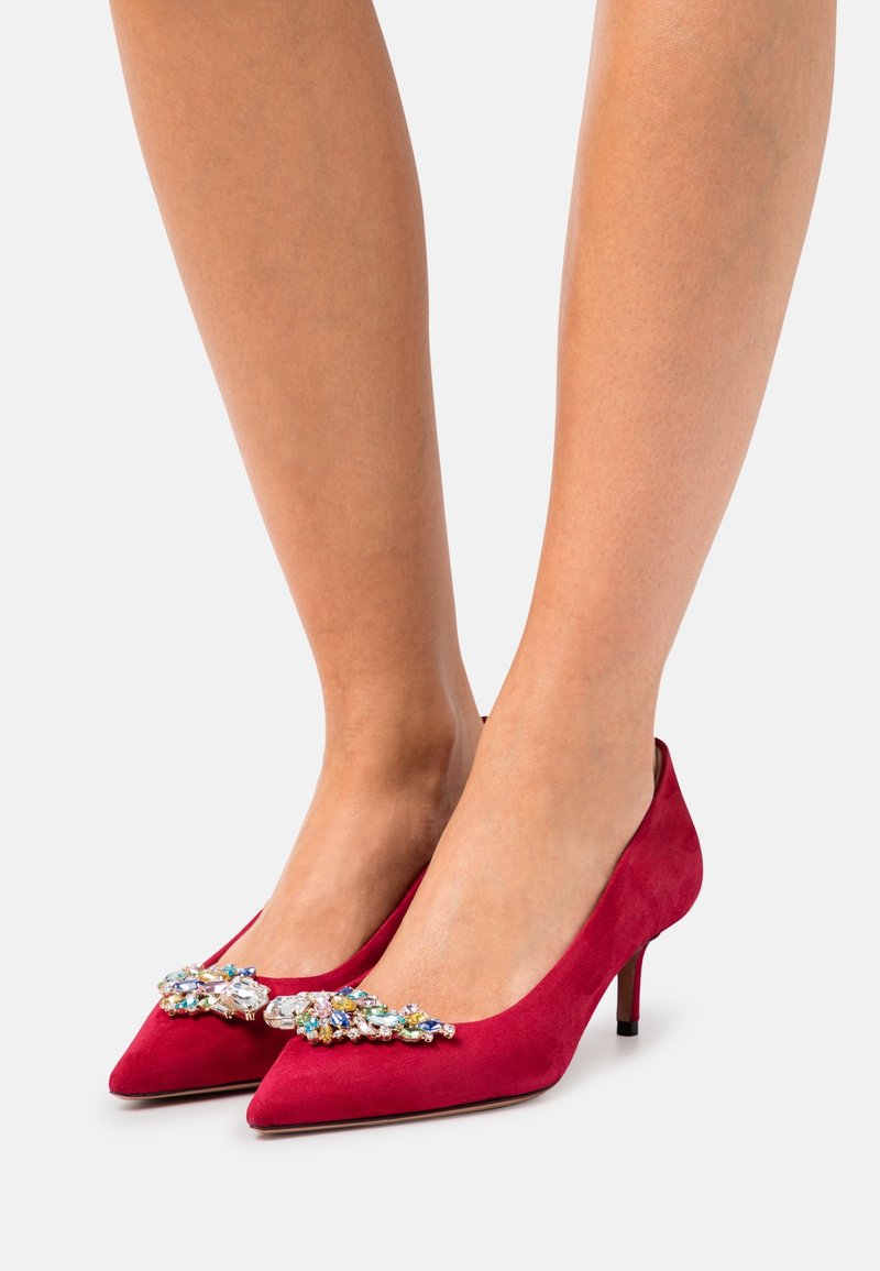 Czerwone zamszowe szpilki z ostrym czubkiem oraz zdobieniami z kryształów z przodu. Posiadają smukły obcas, co dodaje elegancji.