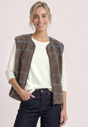 Junge Frau lächelt, trägt eine braune karierte Fleeceweste über einem weißen Langarmshirt und dunkle Jeans, vor einem schlichten hellen Hintergrund.