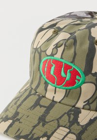 Καπέλο παραλλαγής με μοτίβα σε πράσινο και μπεζ, με κεντρικό κόκκινο οβάλ έμβλημα περιβαλλόμενο από πράσινο και υφές στην επιφάνεια.