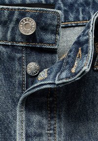 Tela de denim con costuras naranjas, que presenta dos botones a presión de metal y una solapa de botón parcialmente desabrochada.