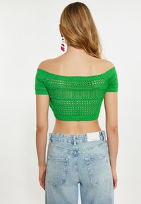 Top scurt tricotat verde, cu un design off-shoulder și modele texturate, asortat cu blugi lejeri cu talie înaltă, de culoare albastru deschis.