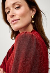 Rote metallische Bluse mit gerafften Puffärmeln und schimmernder Textur. Goldene, abstrakte Ohrringe ergänzen das Design.