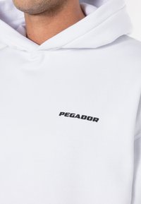 Moletom branco feito de tecido macio, apresentando um logotipo preto "PEGADOR" no peito. O design inclui um capuz com cordão e um bolso canguru.