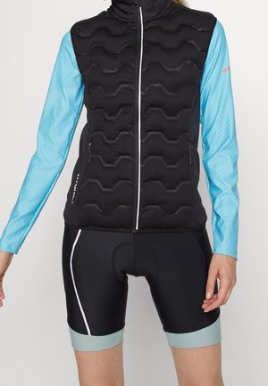 Svart vadderad väst över en ljusblå långärmad topp med geometriska mönster, i kombination med svarta cykelshorts med ljusblå fållar.