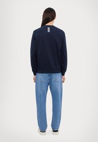 Jūras spēku pelēks džemperis ar garām piedurknēm, apaļu kaklu un baltu svītru detaļu mugurpuss, apvienots ar gaiši ziliem brīvi krītošiem džinsiem.