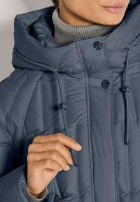 Grijze geïsoleerde jas met een hoge kraag, voorzien van een trekkoordkap, knopen en ribbelstructuur aan de onderkant. Glad materiaal met stikseldetails.