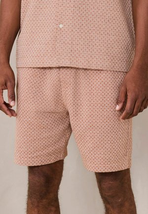 Mann trägt beige strukturierte Strickshorts und ein passendes Hemd mit Knöpfen, steht mit entspannt an den Seiten hängenden Armen vor neutralem Hintergrund.