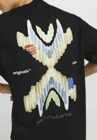 Sort t-shirt med et farverigt abstrakt design på ryggen, i nuancer af grøn, blå og beige, samt hvide accenter. Inkluderer brandtekst.