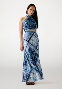 Robe maxi florale bleue avec un col halter à motifs, un ventre nu et une jupe fluide, présentant des nuances variées de bleu et des motifs complexes.