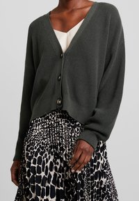 Dunkelgrüner gestrickter Cardigan mit V-Ausschnitt, ausgestattet mit Knöpfen und gerippten Bündchen, kombiniert mit einem Rock im Schwarz-Weiß-Muster.