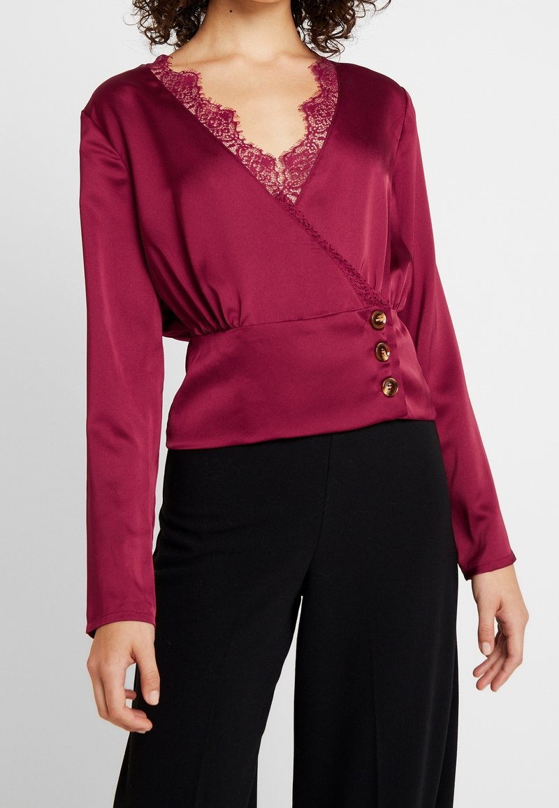 Femme portant un chemisier cache-cœur bordeaux à manches longues avec bordure en dentelle et trois boutons à la taille, associé à un pantalon noir.