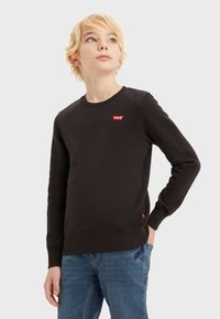 Sudadera negra de mezcla de algodón, con cuello redondo, puños acanalados y un parche del logo rojo de Levi's en el pecho. Combinada con jeans azules.