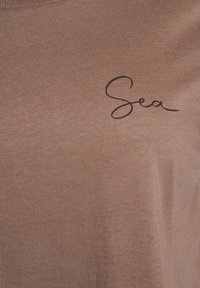 Bruine katoenen T-shirt met een donker cursief "Sea" ontwerp aan de voorkant. Gladde textuur en korte mouwen met een ronde hals.