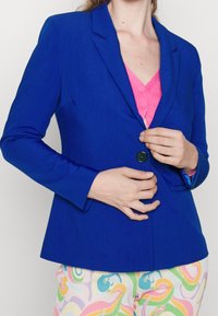 Modrý přizpůsobený blejzr s jedním černým knoflíkem, s klopami, v kombinaci s růžovým topem na knoflíky a vzorovanými multicolor kalhotami.