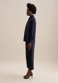 Blusa blu navy con colletto e maniche a palloncino, abbinata a pantaloni blu navy a vita corta e tacchi neri con cinturini. Tessuto di superficie liscia.