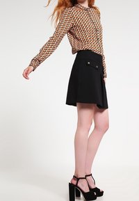 Langärmlige gemusterte Bluse in Orange, Braun und Creme; schwarze A-Linien-Rock mit Fronttaschen; schwarze High-Heelsandalen mit Knöchelriemen.