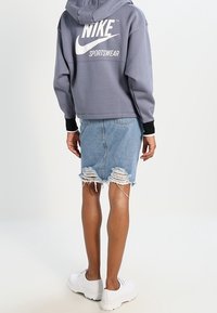 Lila kort hoodie med vitt "Nike Sportswear"-logotyp, tillsammans med en sliten jeanskjol med fransad fåll och vita chunky sneakers.