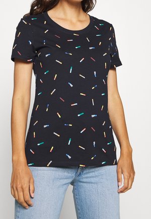 Femme portant un t-shirt noir à manches courtes avec de petits motifs rectangulaires multicolores et un jean bleu clair.