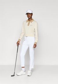 Kremfarget kortermet genser med en beige krage og knappdetaljer, kombinert med hvite bukser og hvite joggesko. Holder en golfklubbe.