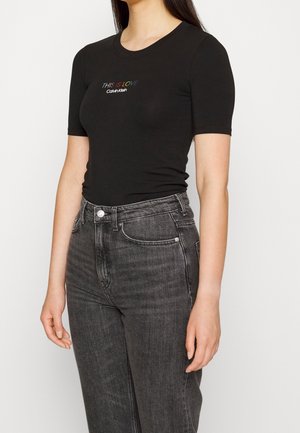 Kobieta w czarnym t-shircie Calvin Klein z "This is love" w kolorach tęczy i ciemnoszarych, wysokich jeansach, stojąca na białym tle.