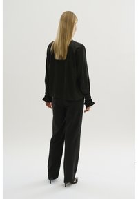 Schwarze Satin-Bluse mit langen Ärmeln und gerafften Manschetten, kombiniert mit weit geschnittenen schwarzen Hosen. Das Modell wird von hinten gezeigt.
