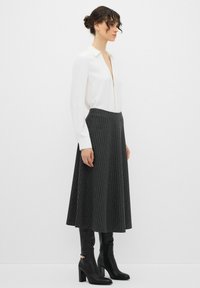 Blusa bianca a maniche lunghe con zip, abbinata a una gonna plissettata grigio scuro con righe verticali e stivali neri con tacco al ginocchio.