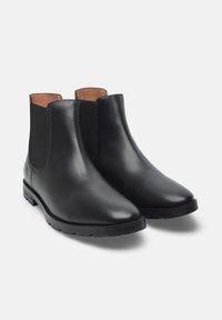 Stivali alla caviglia in pelle nera con pannelli elastici laterali, punta appuntita e suola testurizzata. Finitura liscia con una fodera interna in contrasto color cammello.