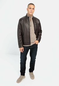Brun læderjakke med quiltede skuldre over en lys beige sweater, parret med mørke jeans og lyse grå sneakers. Ensfarvet baggrund.