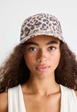 Femme aux cheveux bouclés portant un haut blanc sans manches et une casquette de baseball beige et marron à imprimé léopard, faisant face à l’avant sur un fond uni.
