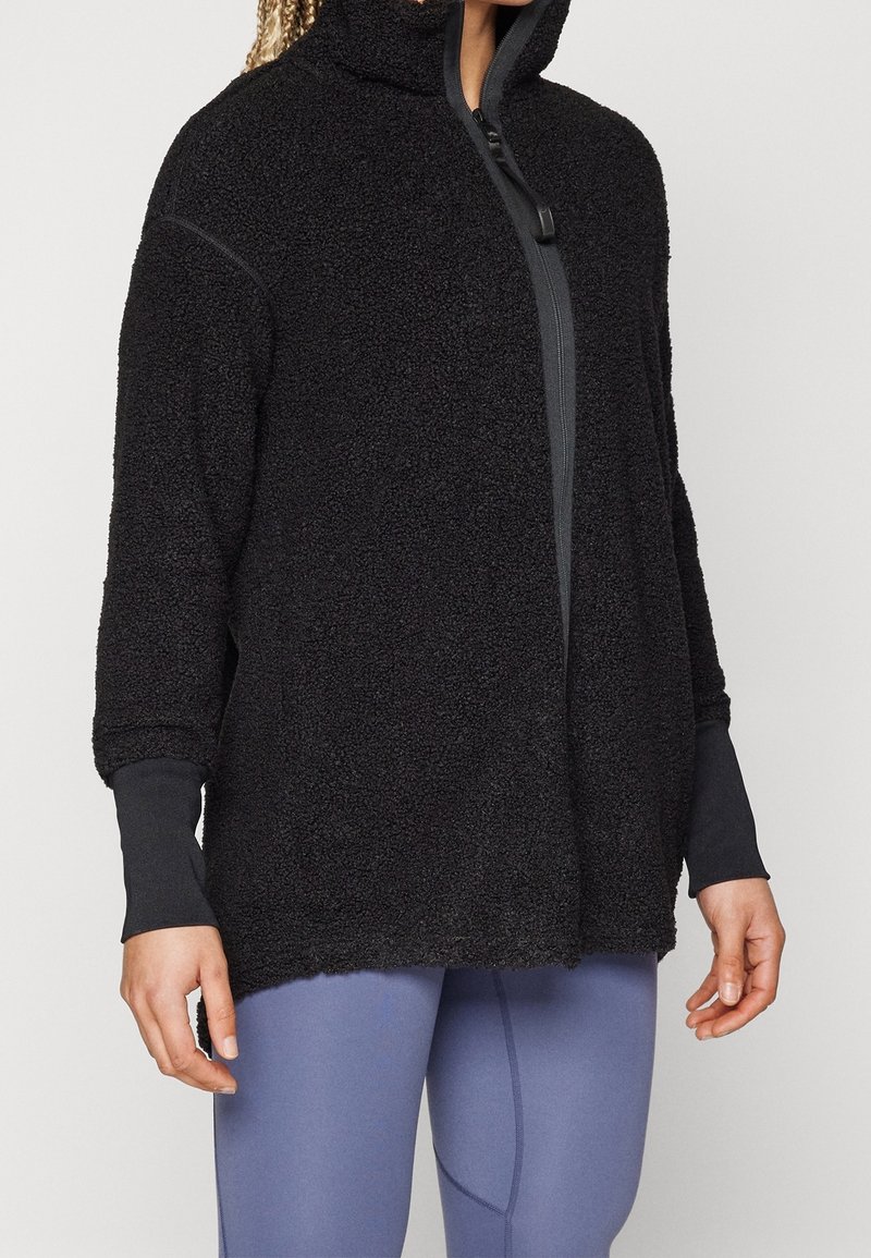 Femme portant une veste noire texturée zippée avec des manches à trou pour le pouce et des leggings bleus ajustés.