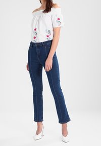Donna che indossa una blusa bianca scollata con ricami floreali, jeans blu aderenti con cuciture frontali e scarpe con tacco alto bianche.