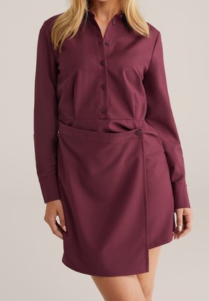 Kobieta nosząca bordową sukienkę z długim rękawem, zapinaną na guziki, z detalem kopertowej spódnicy i kołnierzem, stojąca na jednolitym tle.