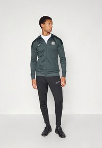 Green zip-up jacket with hood and logo on chest, paired with black sweatpants featuring a white Nike emblem, worn with black footwear.